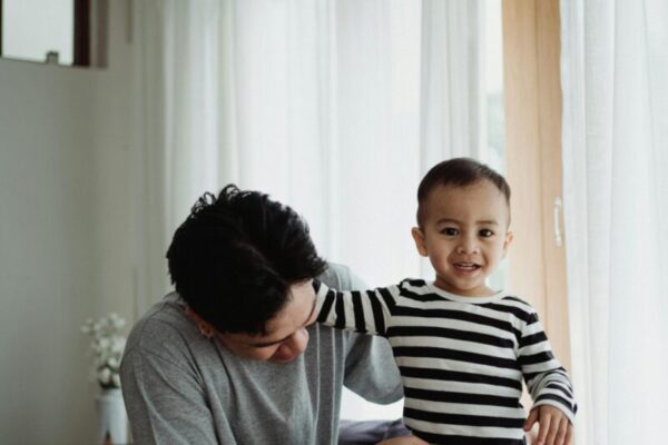 A father and child enjoying a joyful moment indoors. The child smiles happily while standing in a striped shirt.