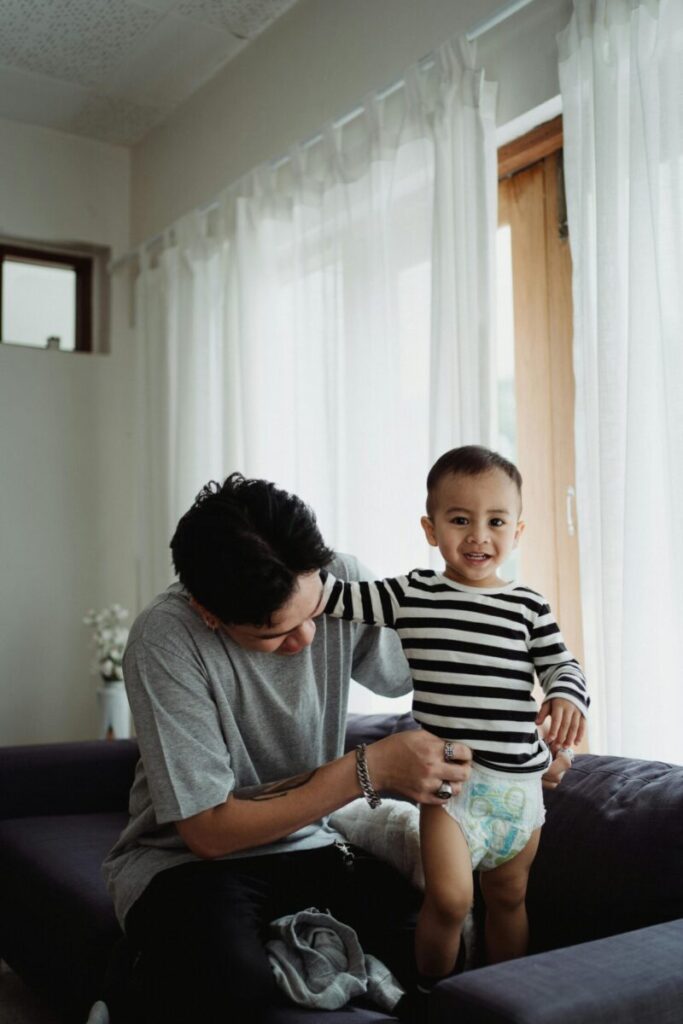A father and child enjoying a joyful moment indoors. The child smiles happily while standing in a striped shirt.